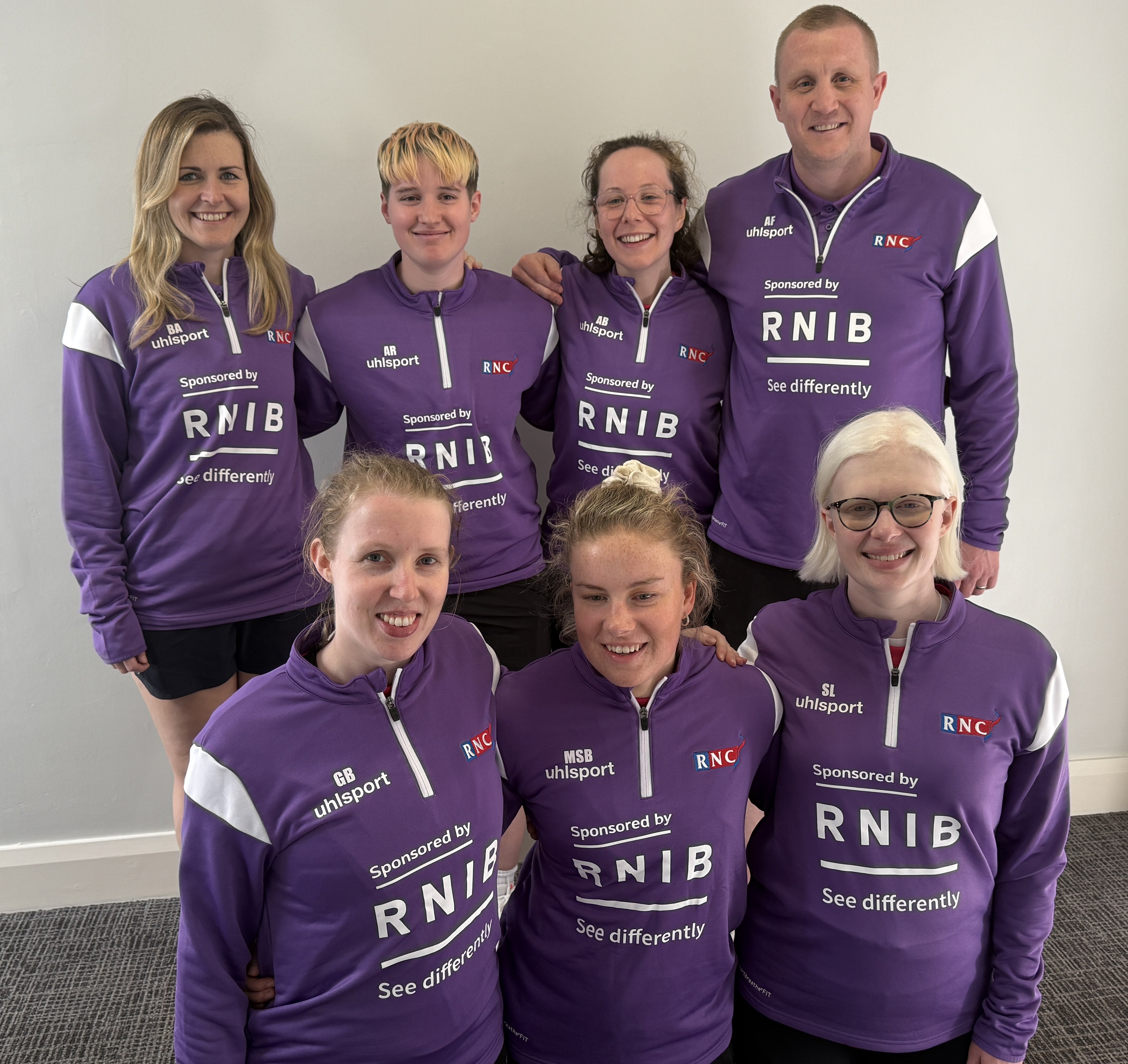the team and coaches pose in their purple off court kit with the RNIB logo in white on it's front