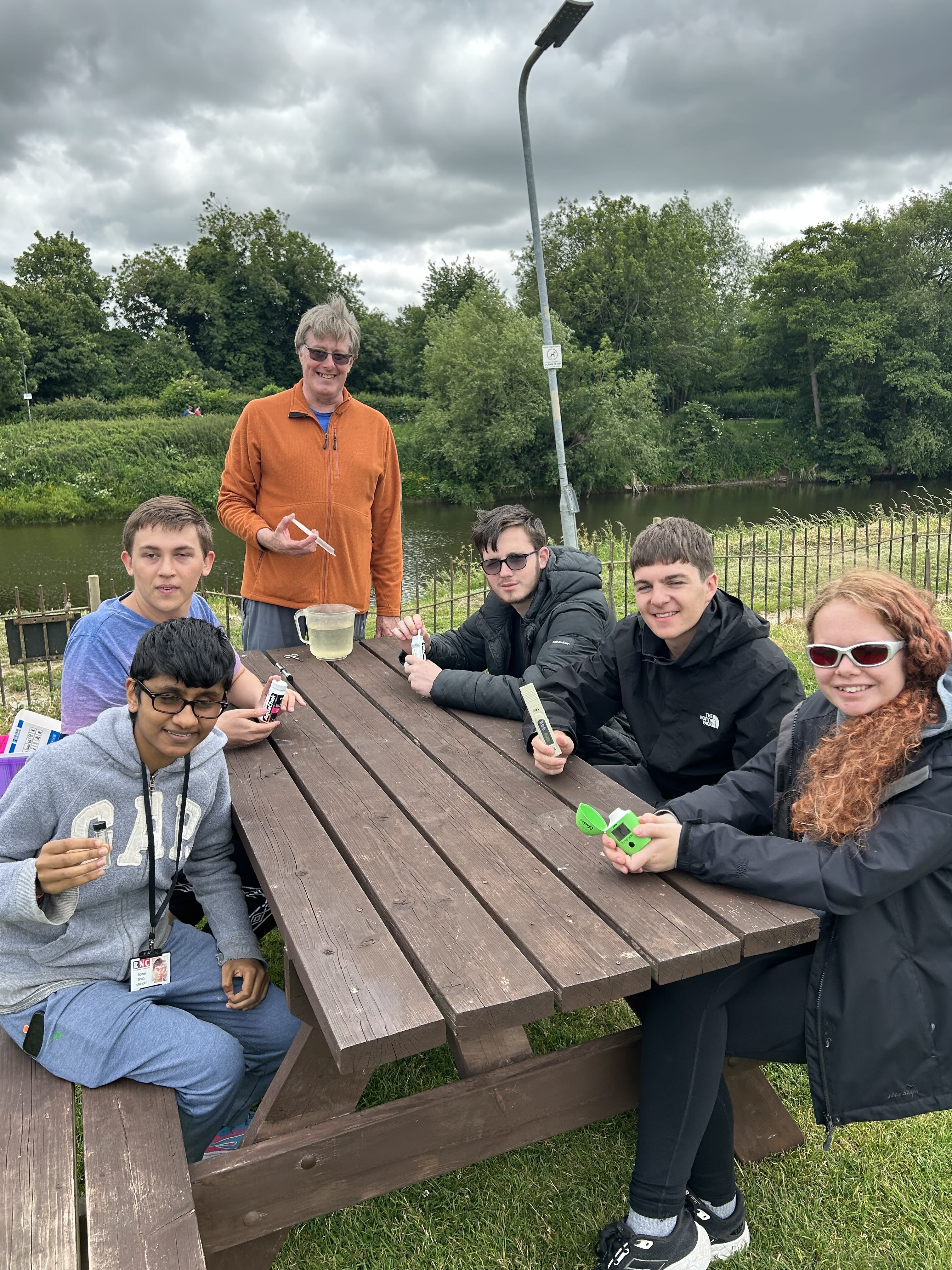 A group of people sitting around a picnic bench next to the River. They are each holding up instruments for testing the water 