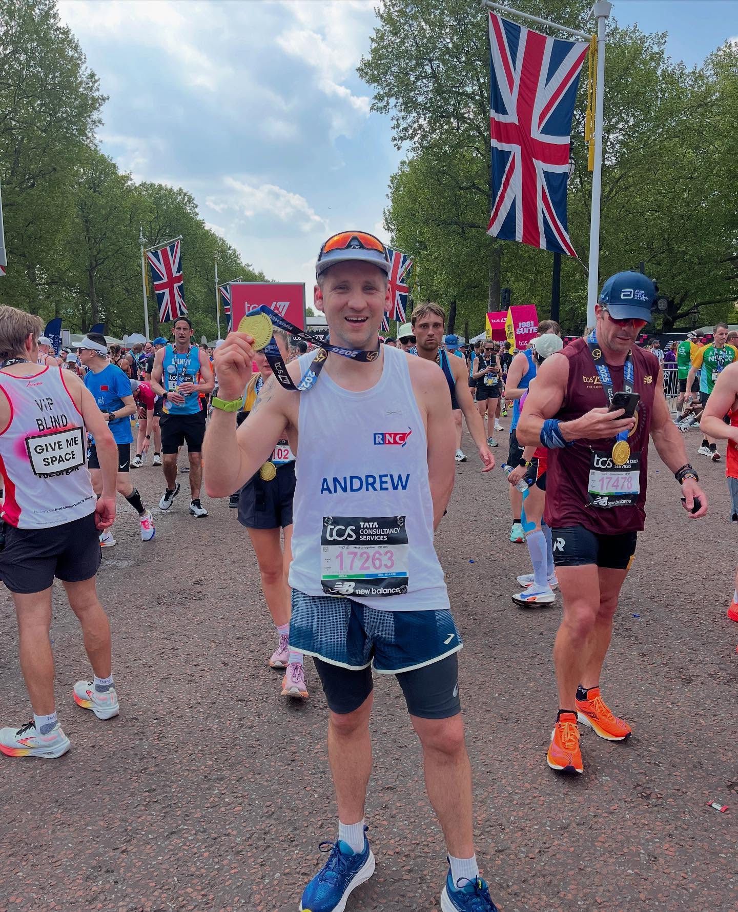 Andrew Davis holds up his medal in the middle of the Mall. He is surrounded by other runners and there are union flags on poles down the length of the road