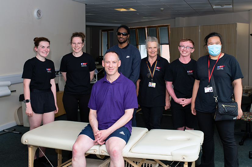 Anthony sits on a massage table with a group of staff and students around him