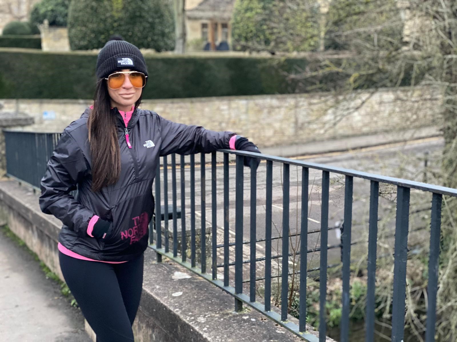 Barbara leans against a fence, she is in training gear a hat and sunglasses ready to start out on a run