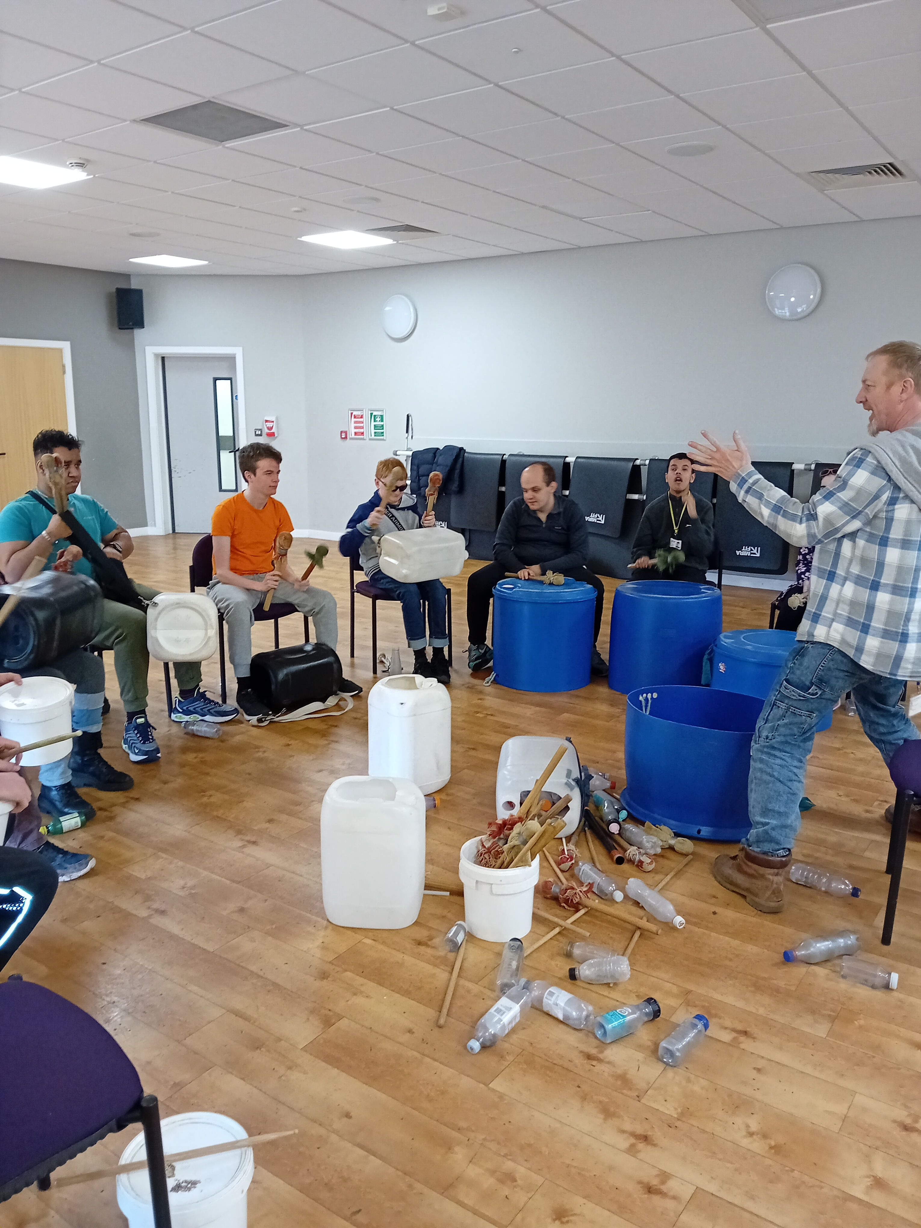 The group sitting in a circle with the upcycled drums in front of them as Bym enthusiastically guides them what to do