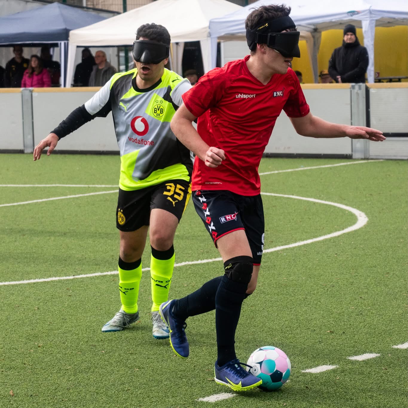 an RNC player in red top, blue shorts and socks on the ball as a Dortmund player tries to catch up