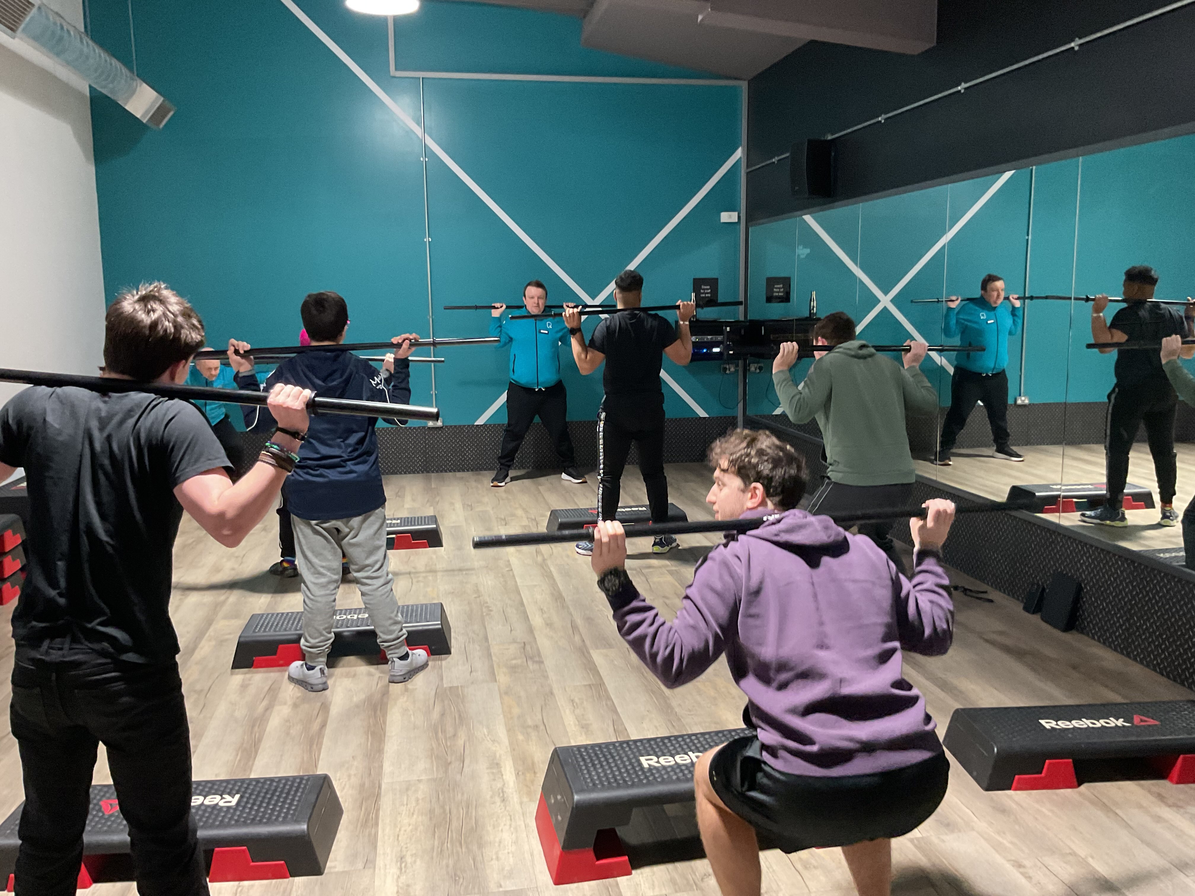 From the back of the studio the students are holding weight bars and are about to squat as Jack leads them through the exercise