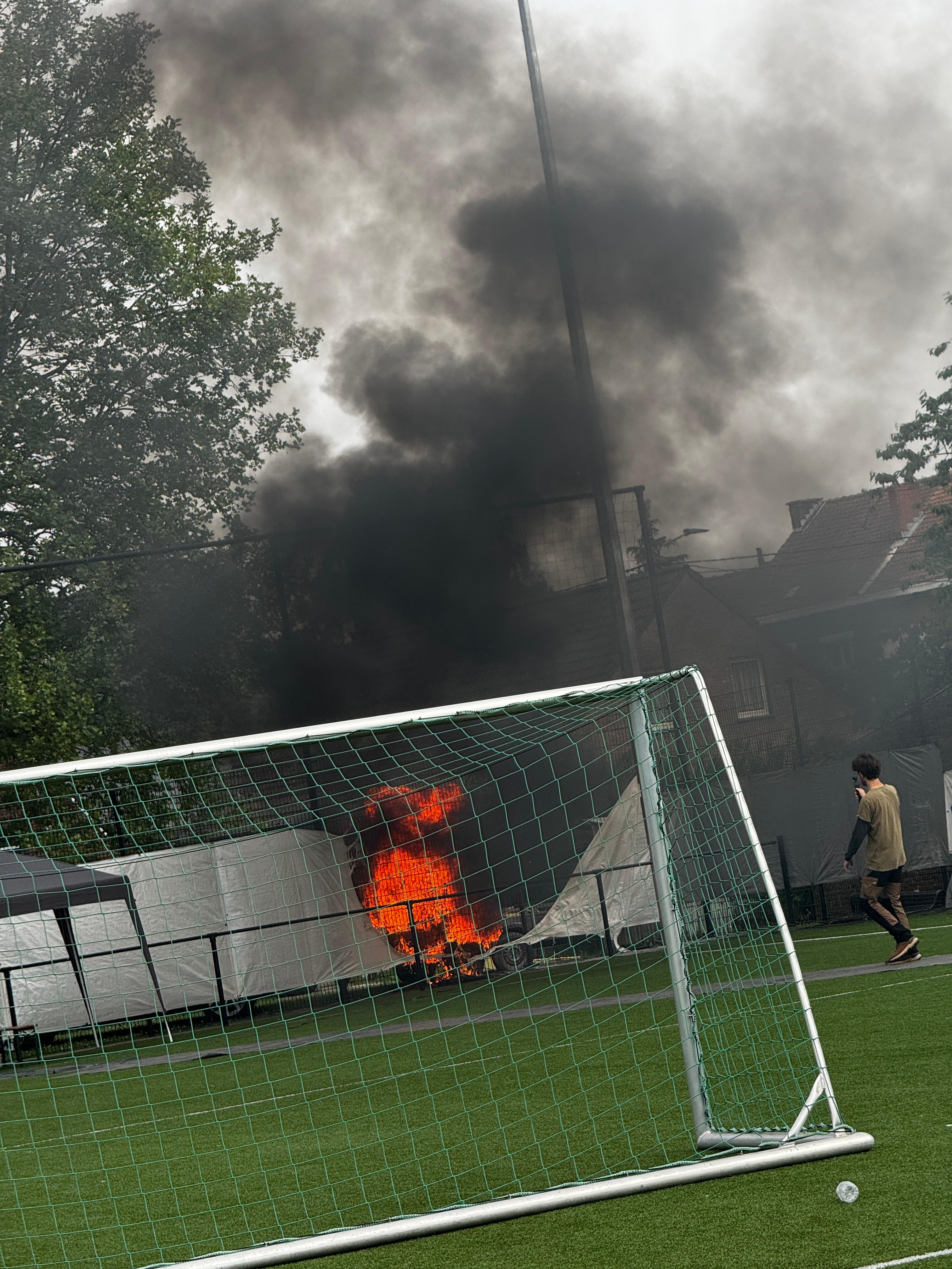 bright orange flames and dark smoke rises into the air on the far side of the pitch behind the boards. A goal is in the foreground