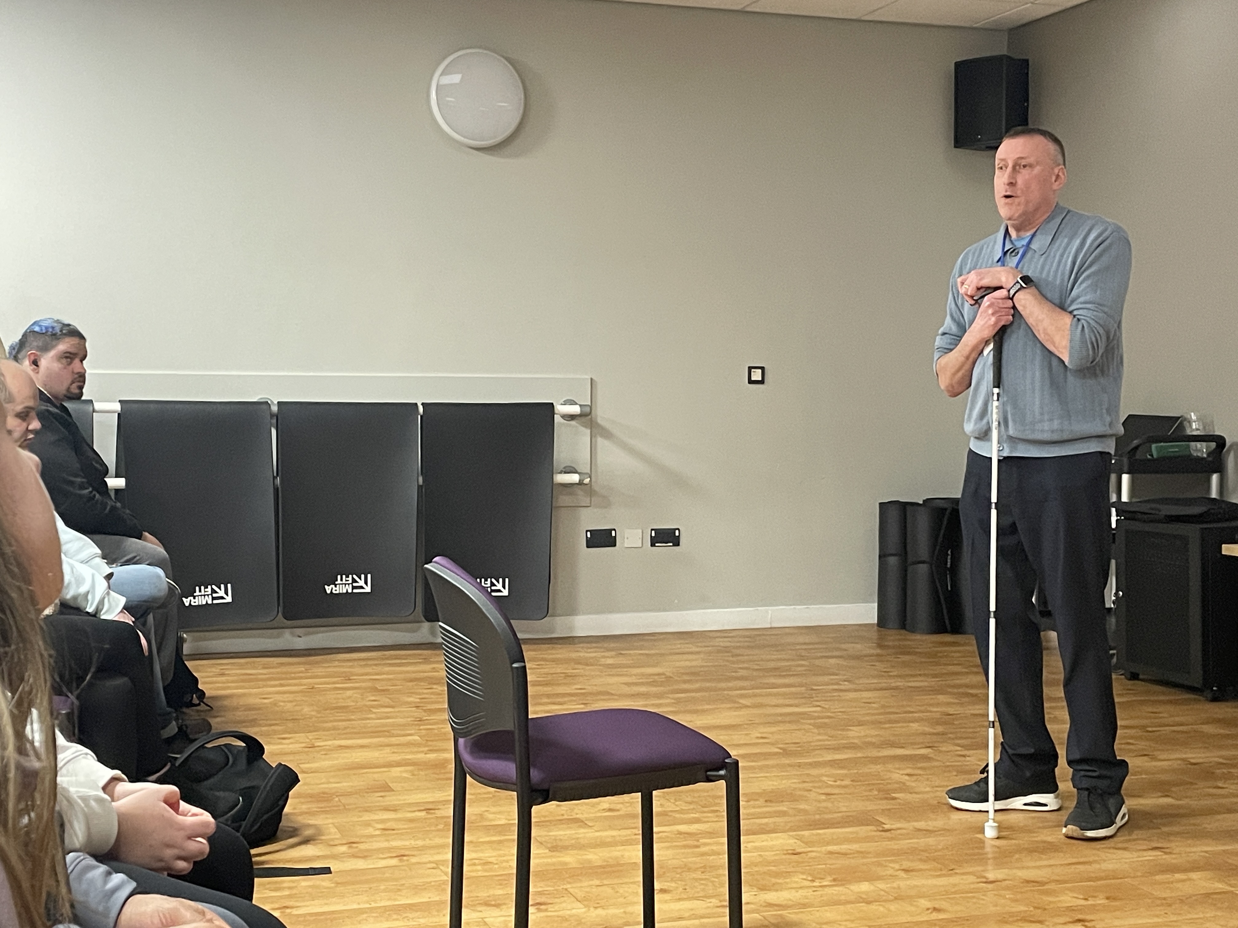 Barry stands at the front of the room facing his audience. He is leaning on his cane