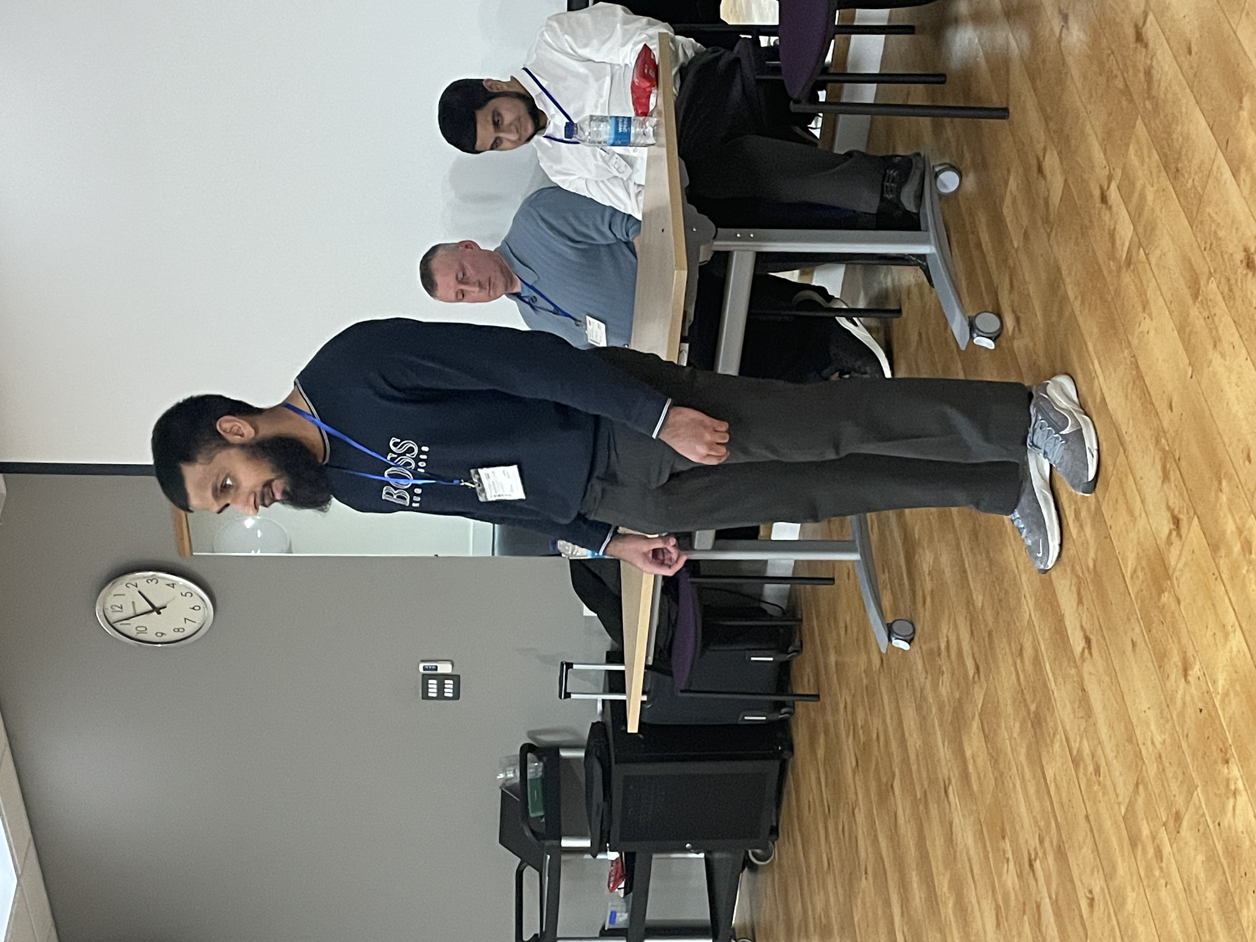 Eesa stands at the front of the room facing his audience. Barry and Mo are sitting at a table behind him.