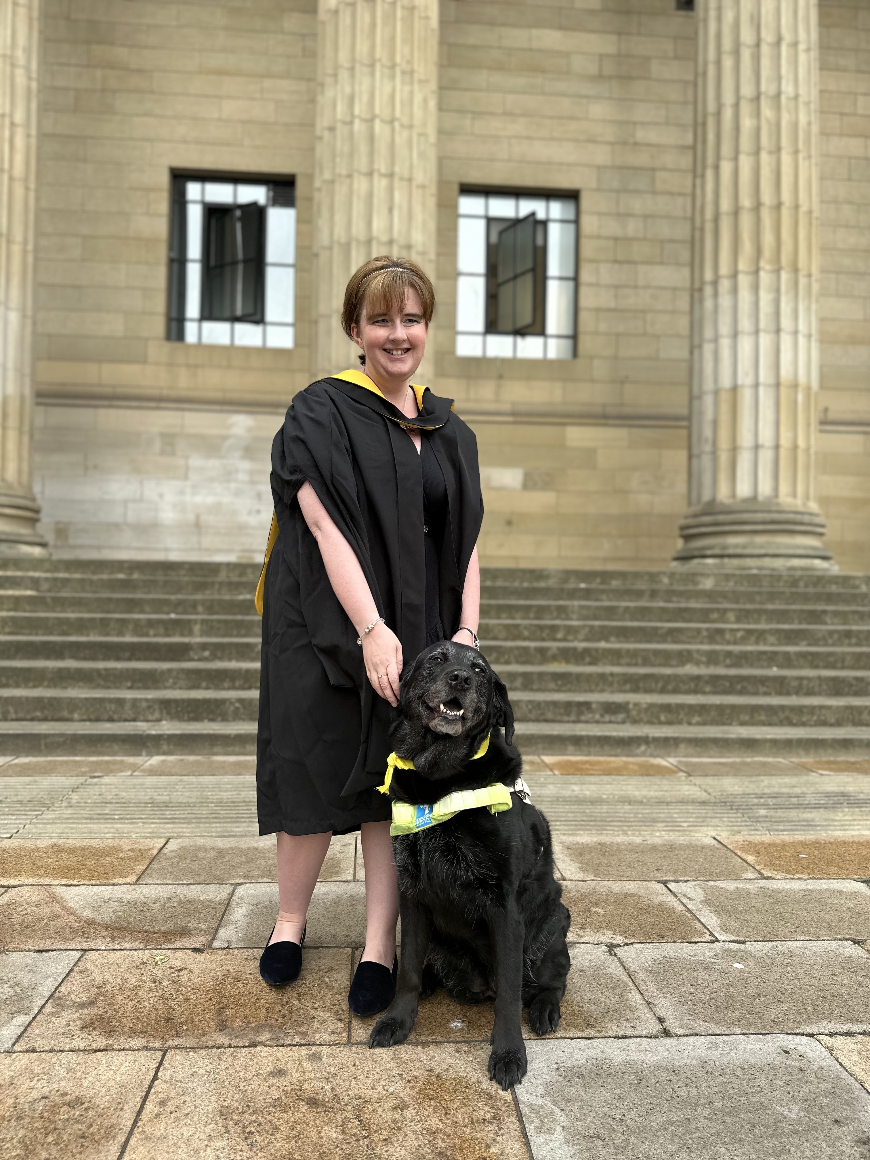 Leanne in her graduation gown with guide dog Wanda who also wears a yellows sash