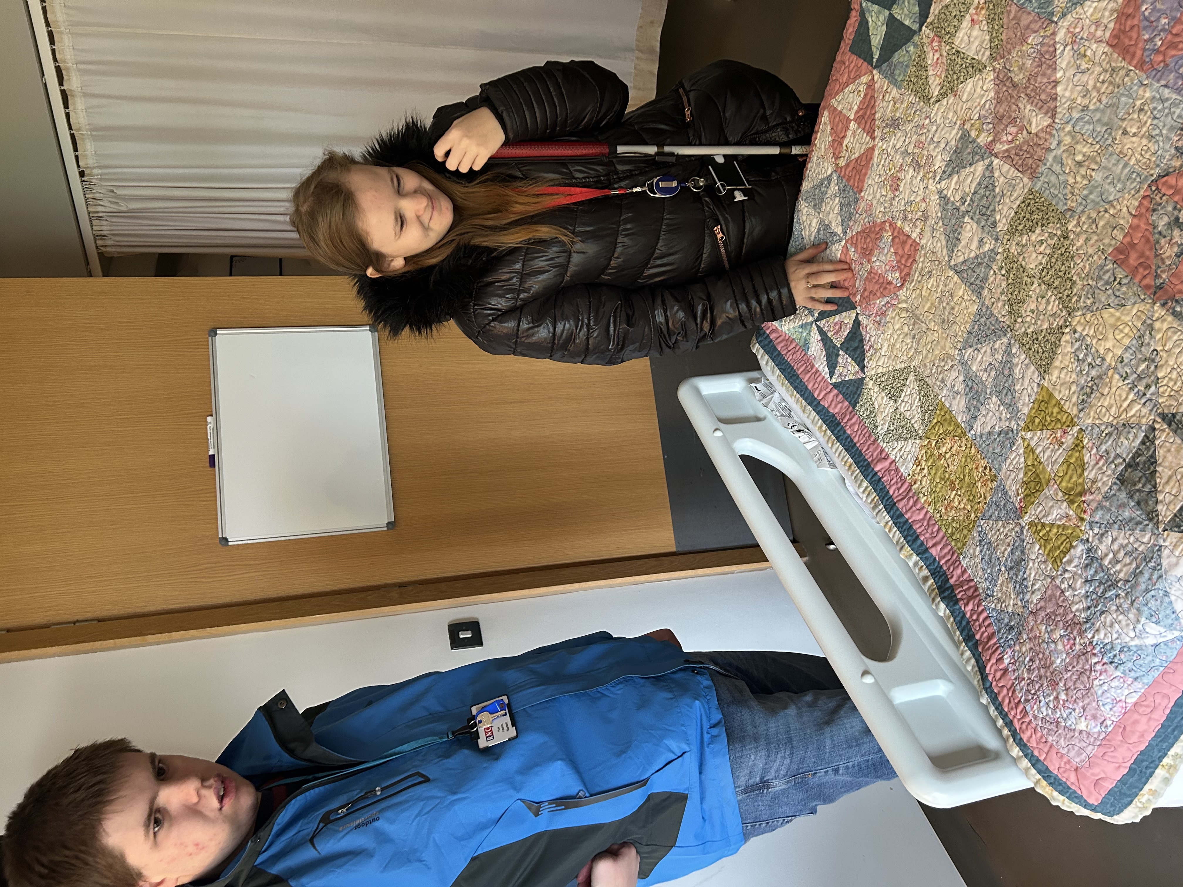 two students stand at the end of an empty hospital bed - one student is exploring the colourful, textured, throw laid across the end of the bed