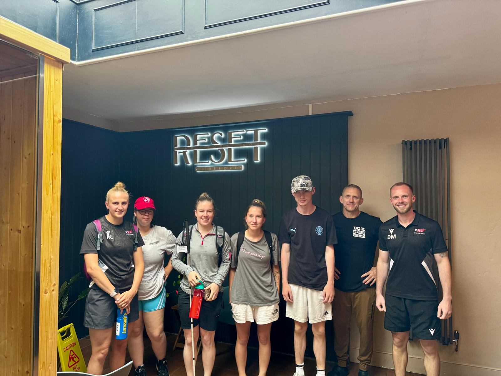 the group of staff and students stand with the RESET owner under the large, lit-up company sign