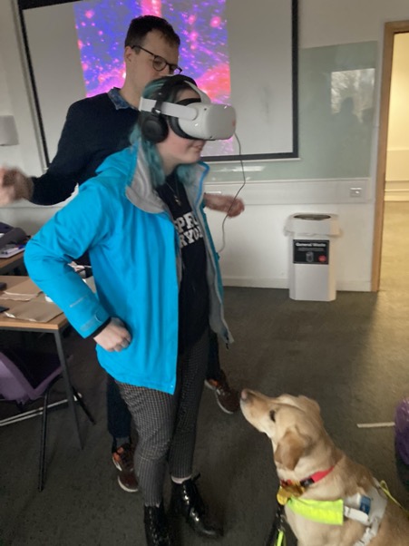 A female student wearing VR goggles with Dr Nic showing how to use them. A guide dog looks on