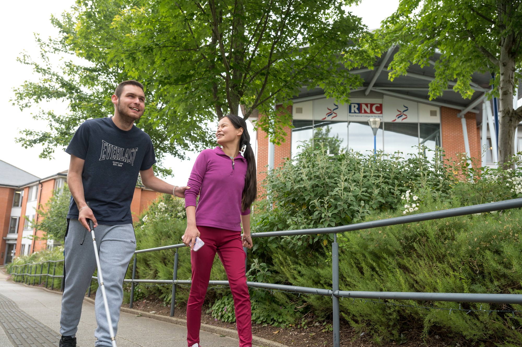 a male student with a cane is guided by a female student walking up from Stephen's hall with the Learning Hub building in the background behind them