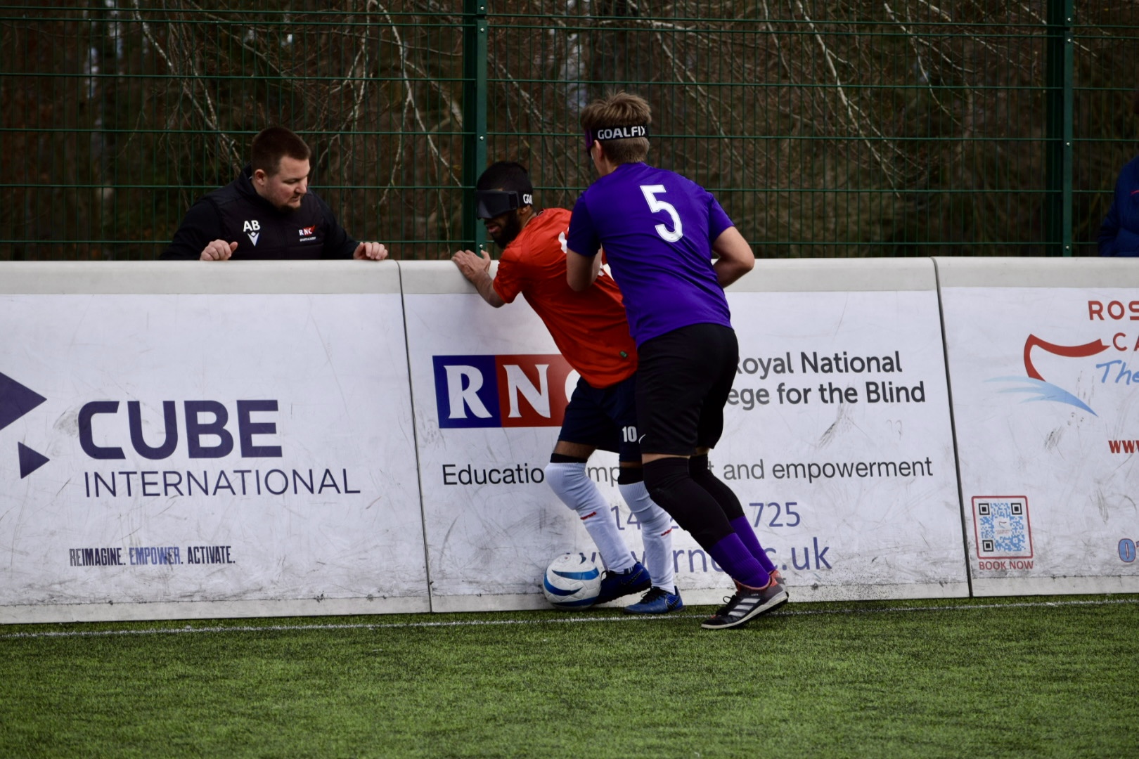 Two blind footballers in action on the pitch. One in a red jersey, the other in a purple jersey. they are both focused on the ball on the ground at their feet. The echo boards with advertisements are behind them