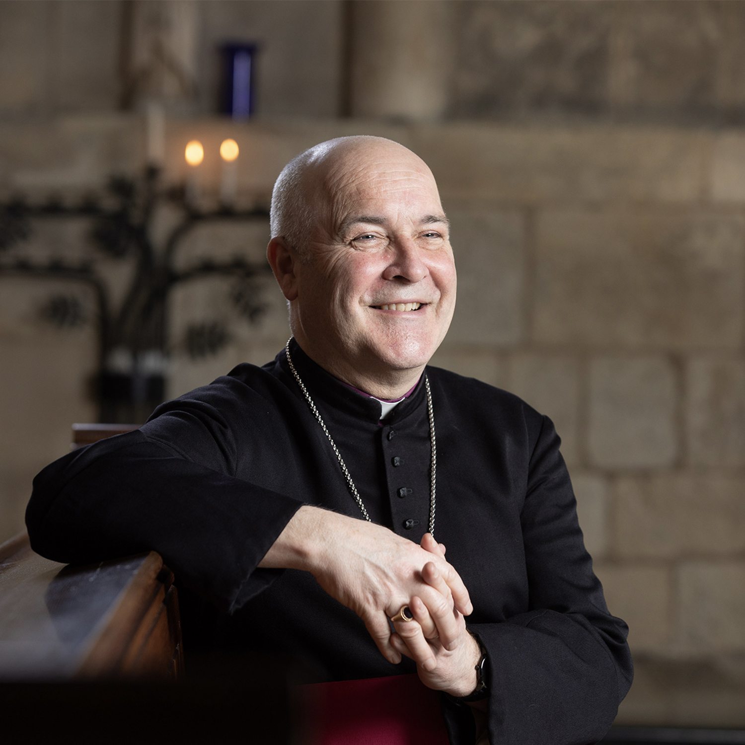 The Archbishop smiles and is in a relaxed seated position in a cathedral setting