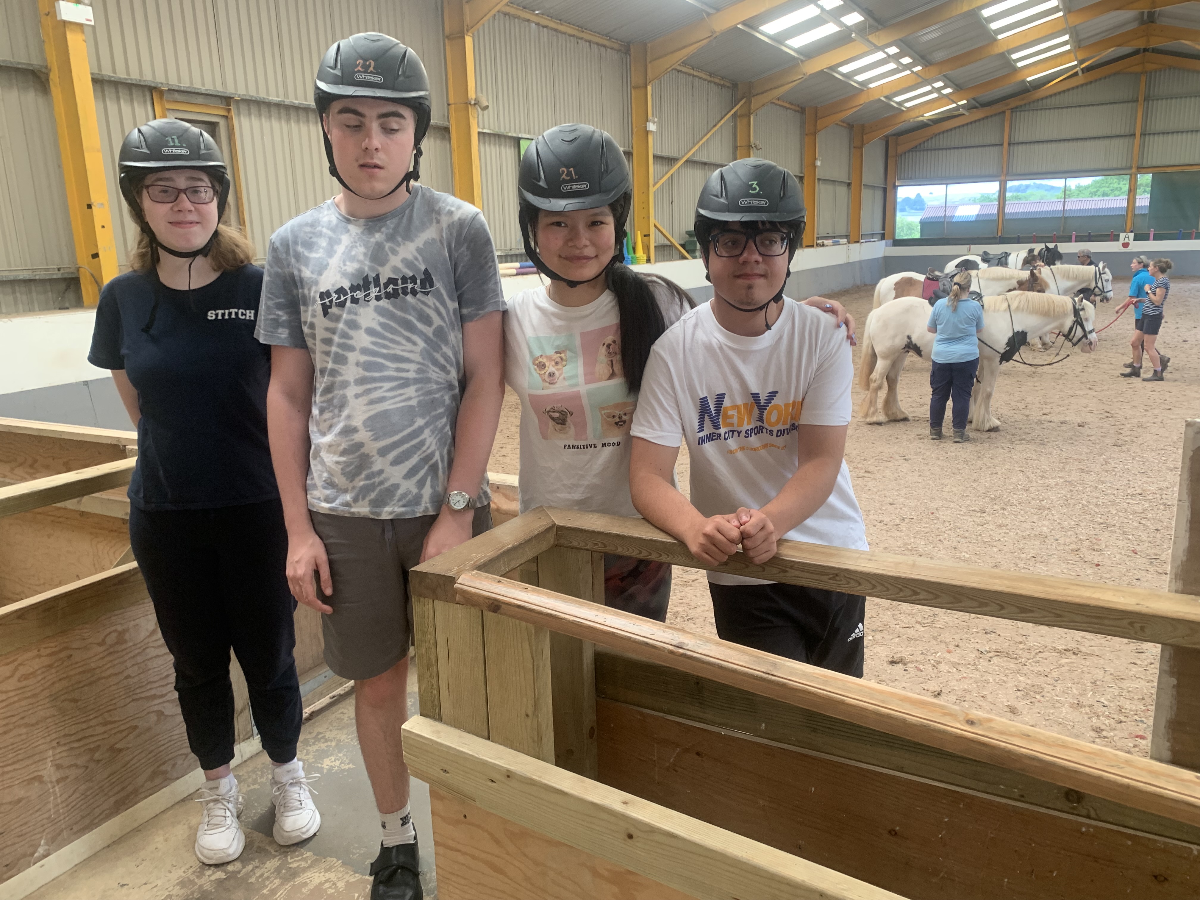 four students wearing riding helmets stand on the edge of the arena waiting for their turn to ride the horses in the background