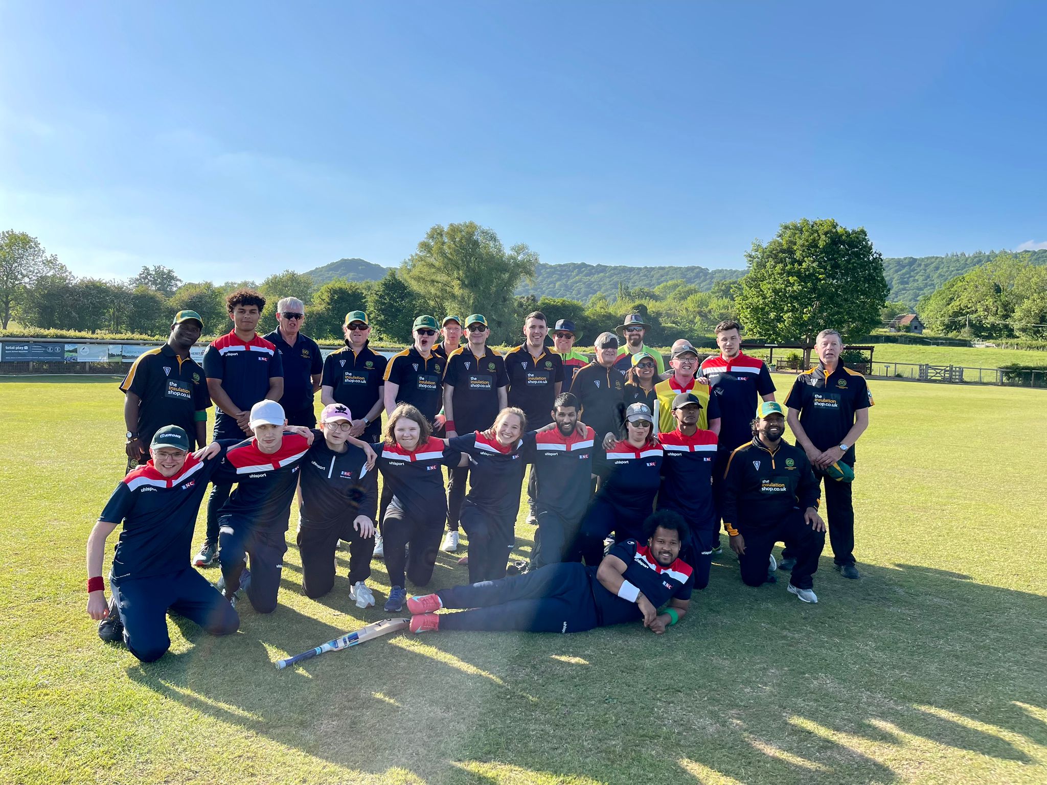 The players from both teams in a group on the pitch. Its a beautiful day with blue sky and open countryside surrounding the pitch